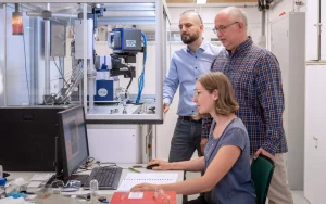 Das Foto zeigt Dr. Janio Venturini (links), Prof. Tom Nilges und Erstautorin Anna Vogel vor einem Messgerät zur Bestimmung von Kristallstrukturen, einem sogenannten Einkristalldiffraktometer.